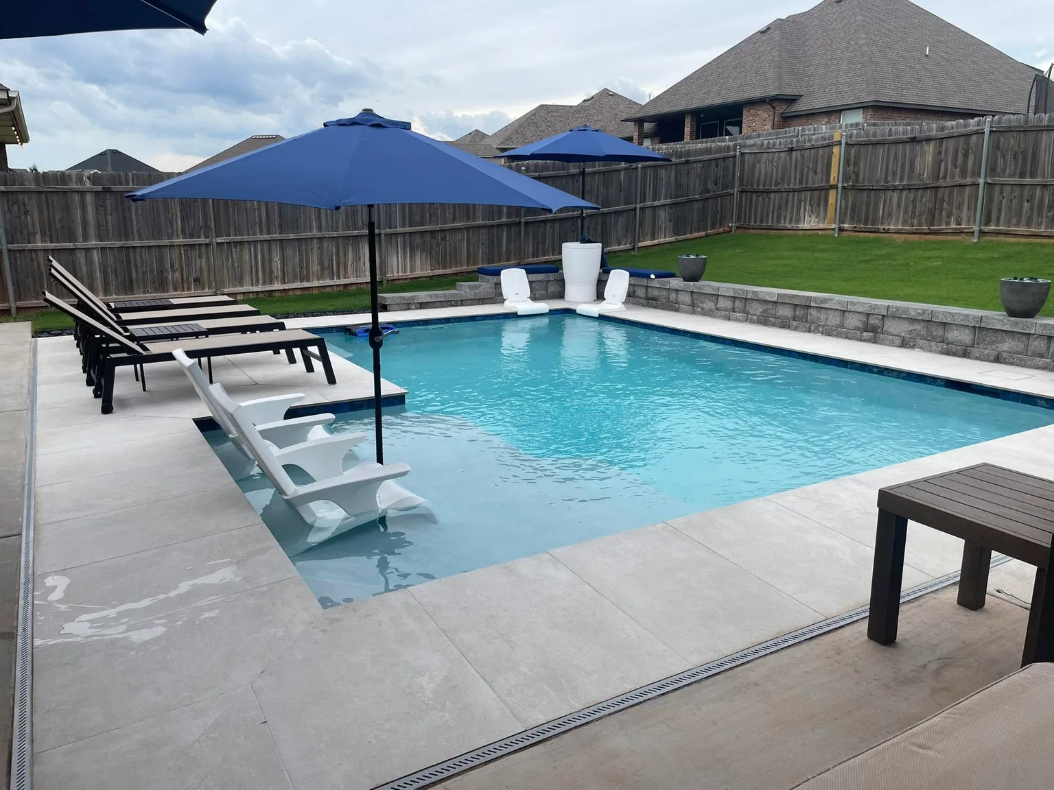 Backyard pool with blue umbrellas, pale coping, and brick homes beyond a wood fence