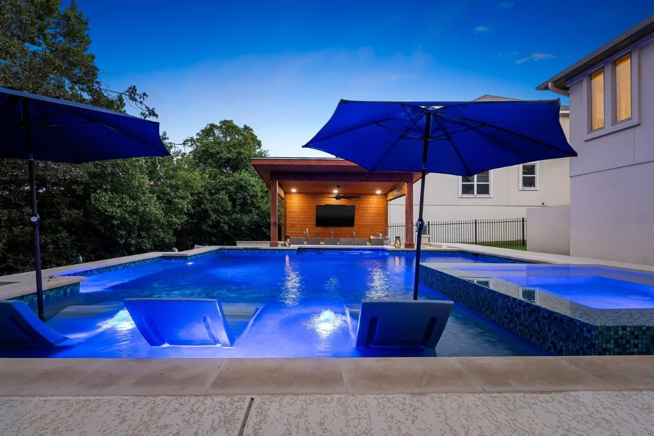 Blue underwater lights illuminate a pool, raised spa, and shallow lounge shelf at dusk